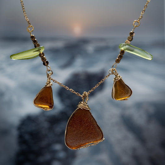 Handmade sea glass necklace featuring warm amber and soft green glass pieces on a gold chain, photographed against a misty beach sunset background.